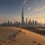 Skyline von Dubai mit dem Burj Khalifa im Hintergrund, Sanddünen und Palmen im Vordergrund bei Sonnenuntergang.
